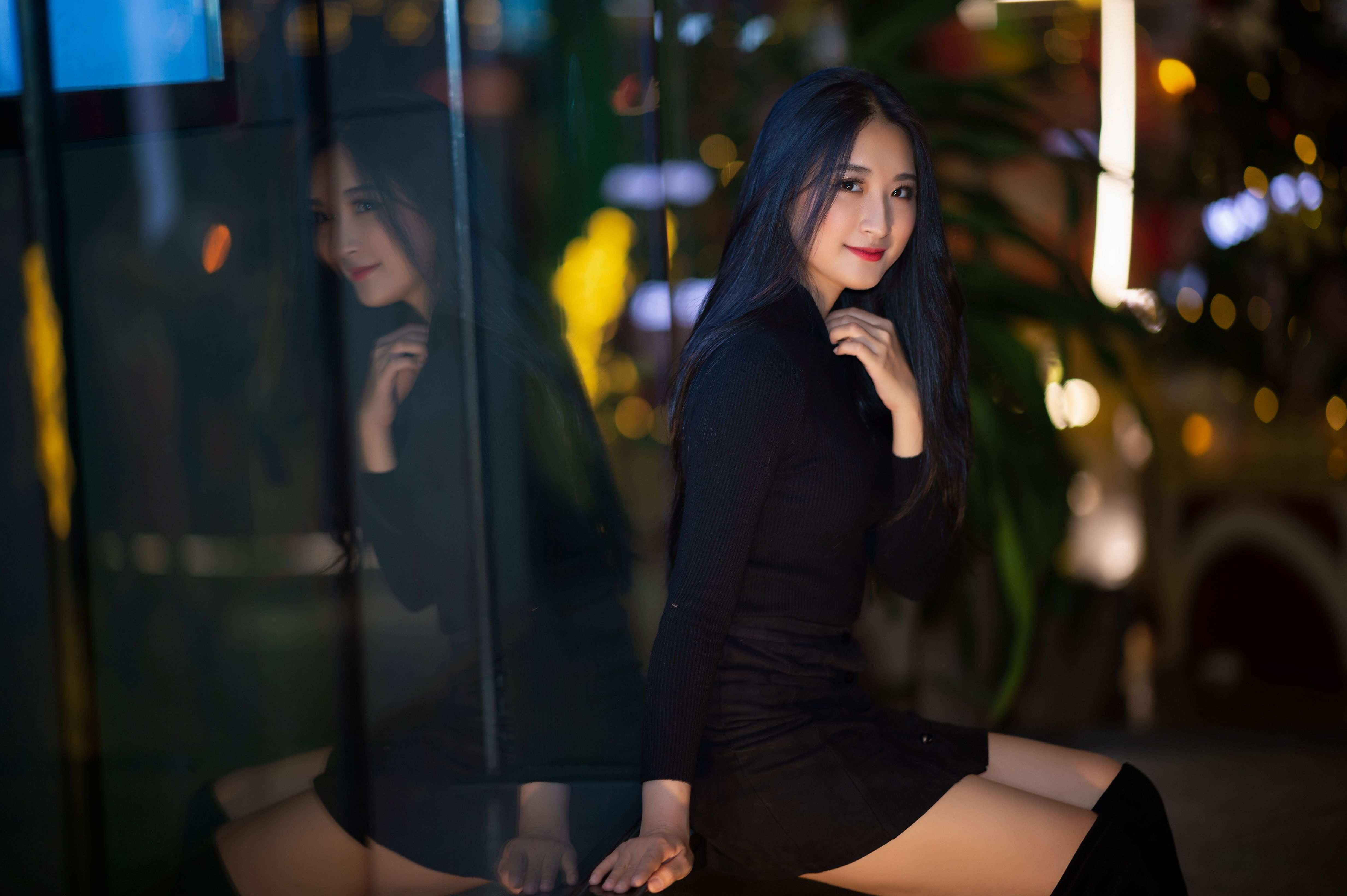 Positive young Asian female in black dress sitting near glass wall with reflection while looking at camera in illuminated town district at night