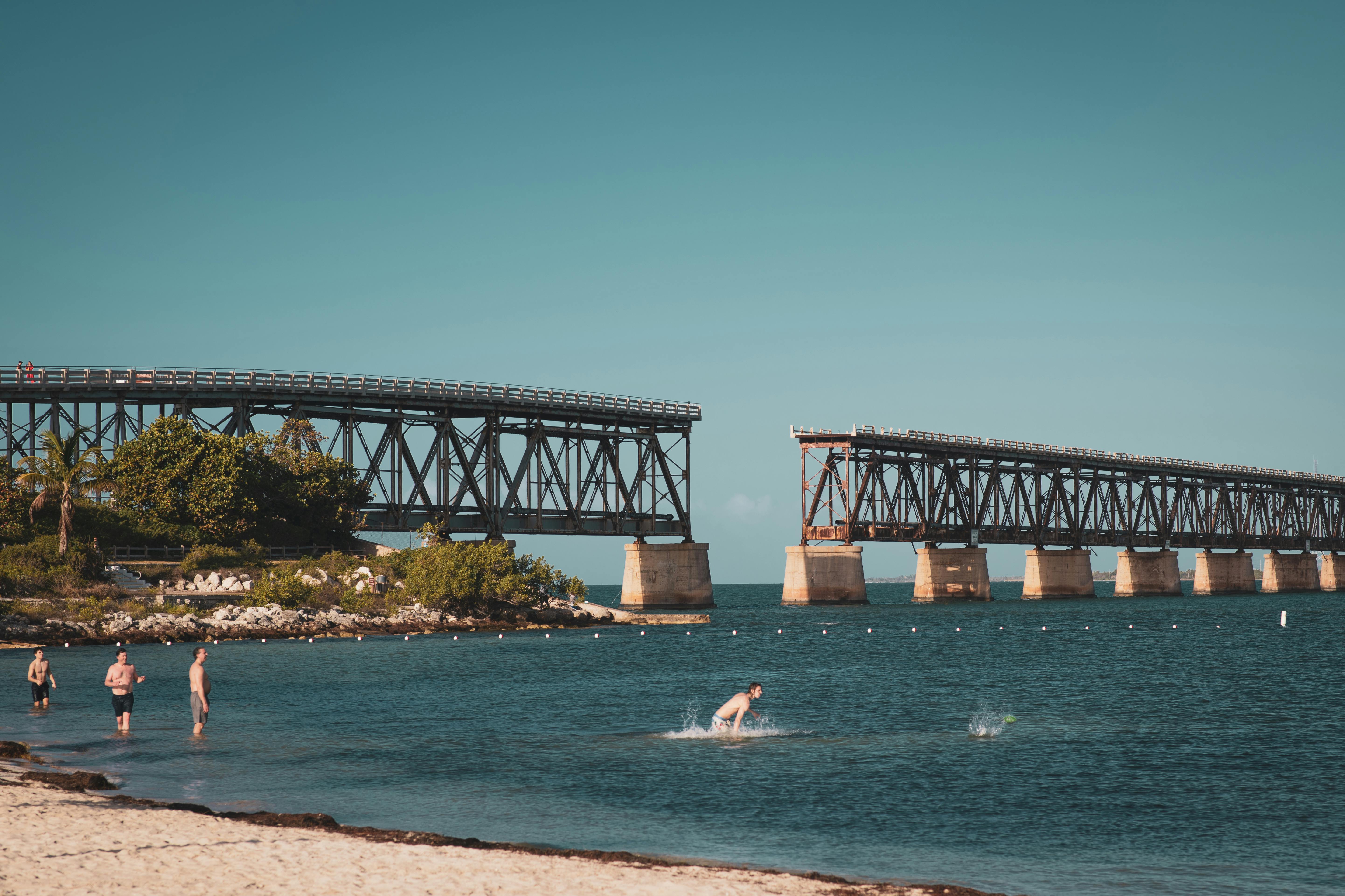 Relaxing beach scene with an old bridge and people swimming under a clear blue sky.