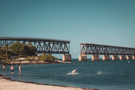 Relaxing beach scene with an old bridge and people swimming under a clear blue sky.