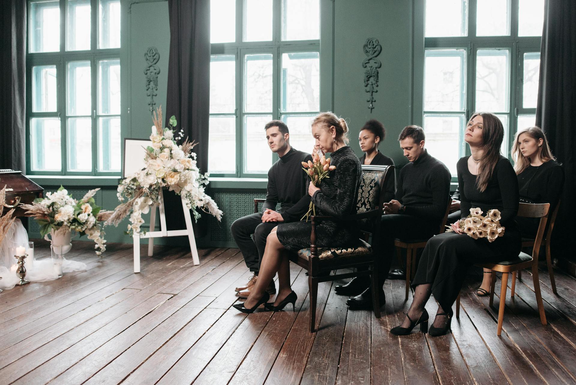 Mourners sitting indoors at a funeral ceremony, dressed in black, holding flowers.