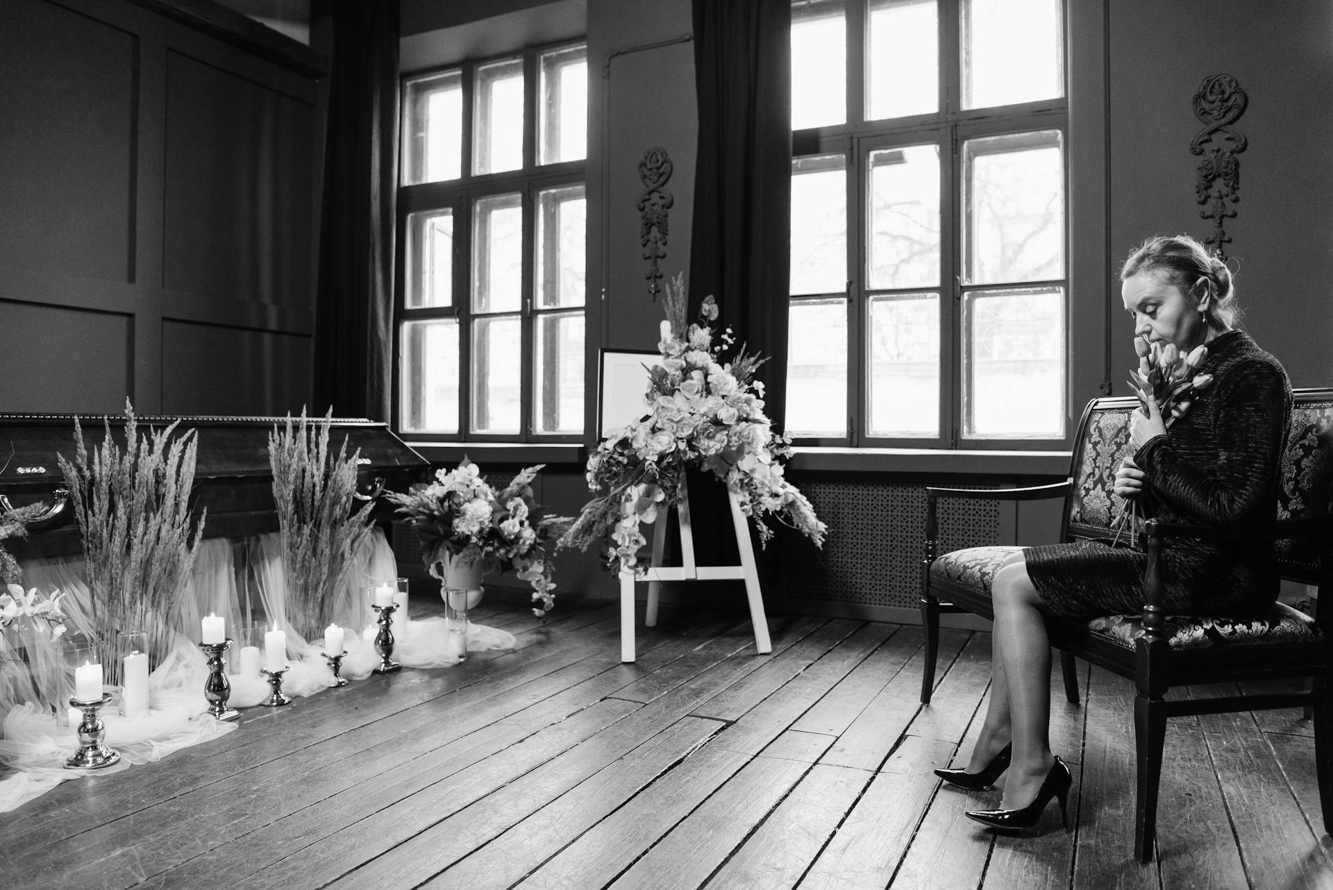 Grayscale funeral scene showing a woman in mourning by a coffin and floral arrangements.