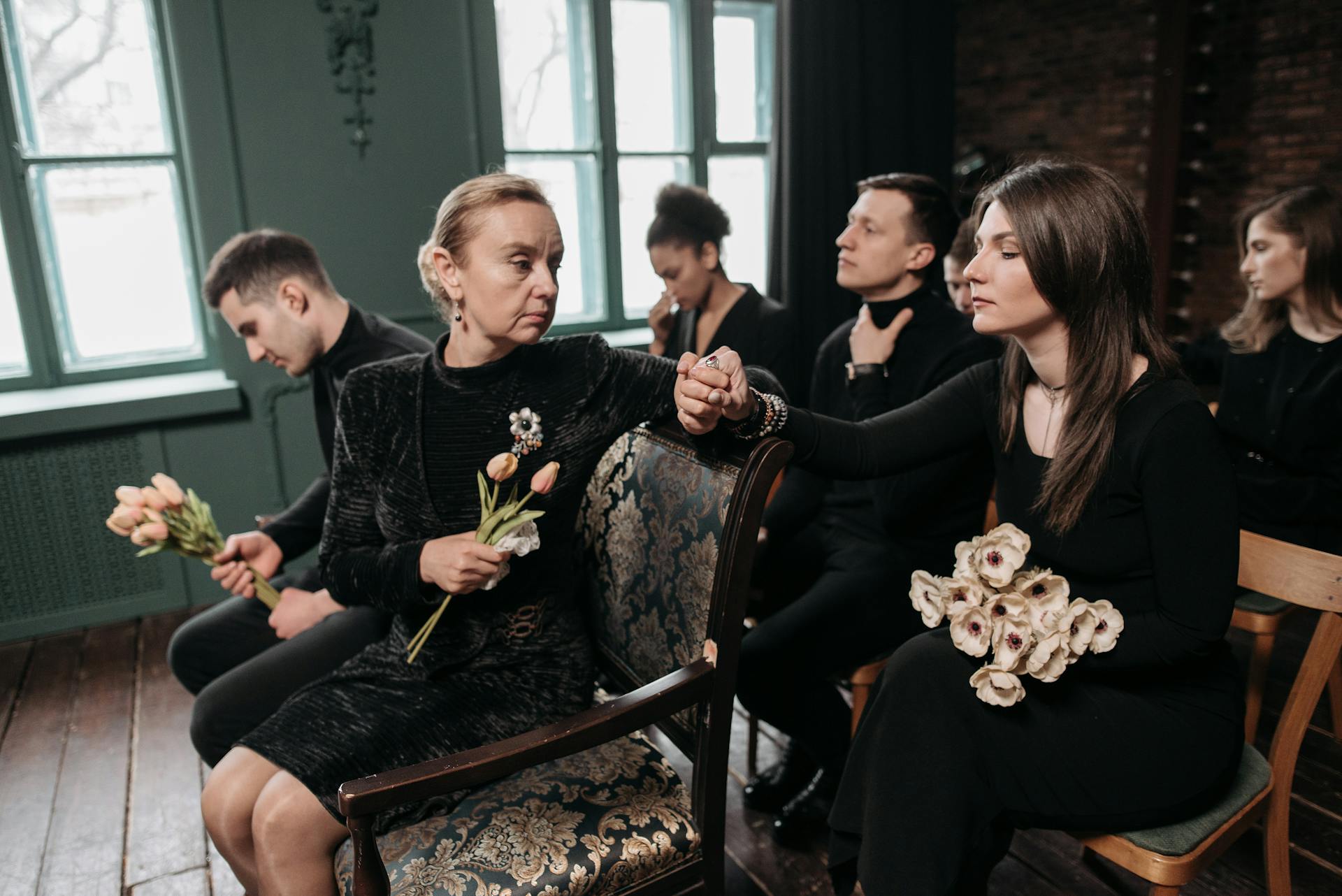 A group of adults dressed in black, holding flowers, at a funeral ceremony indoors, reflecting sorrow.