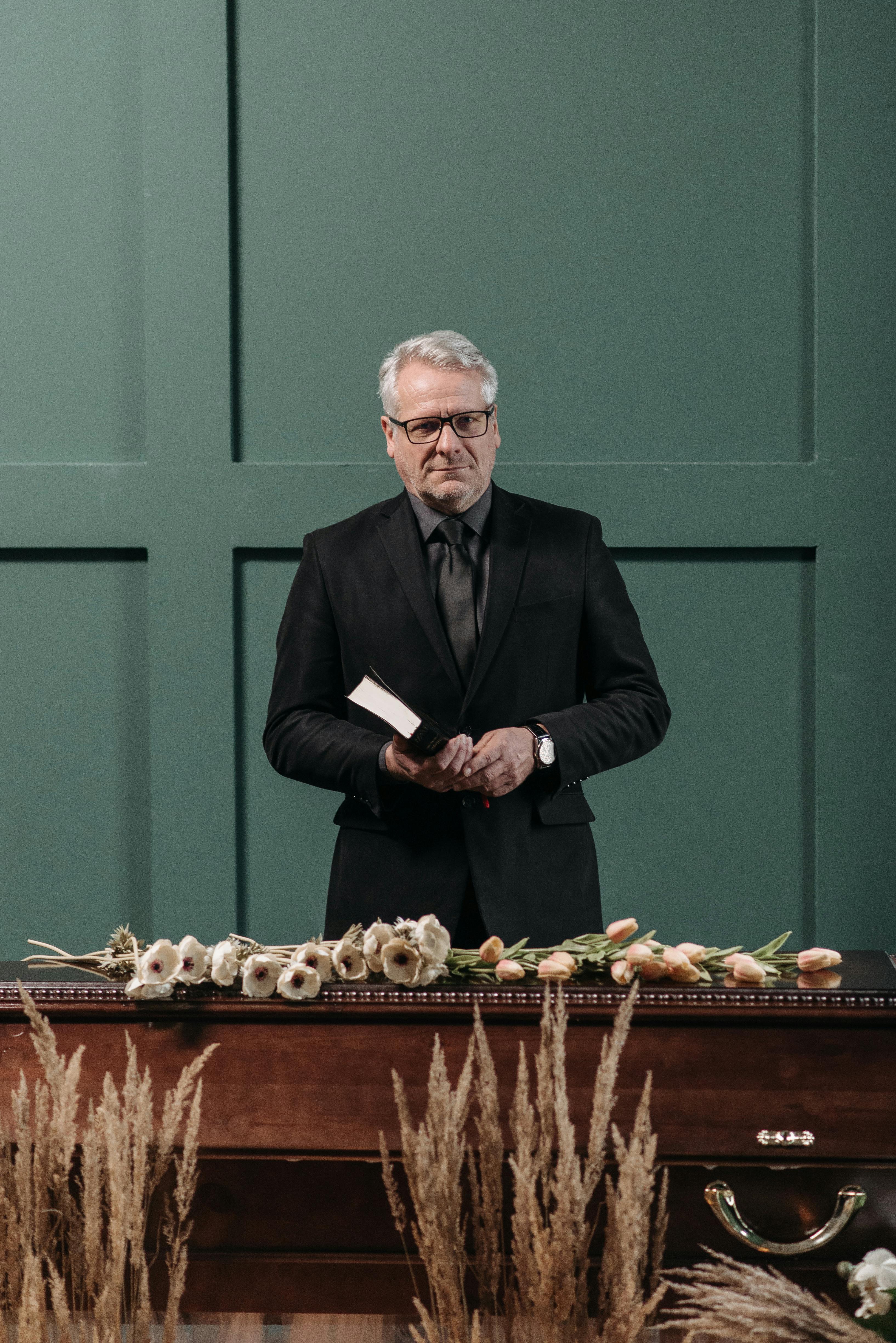 Senior man in a suit holding a book at an elegant indoor funeral setting with flowers.