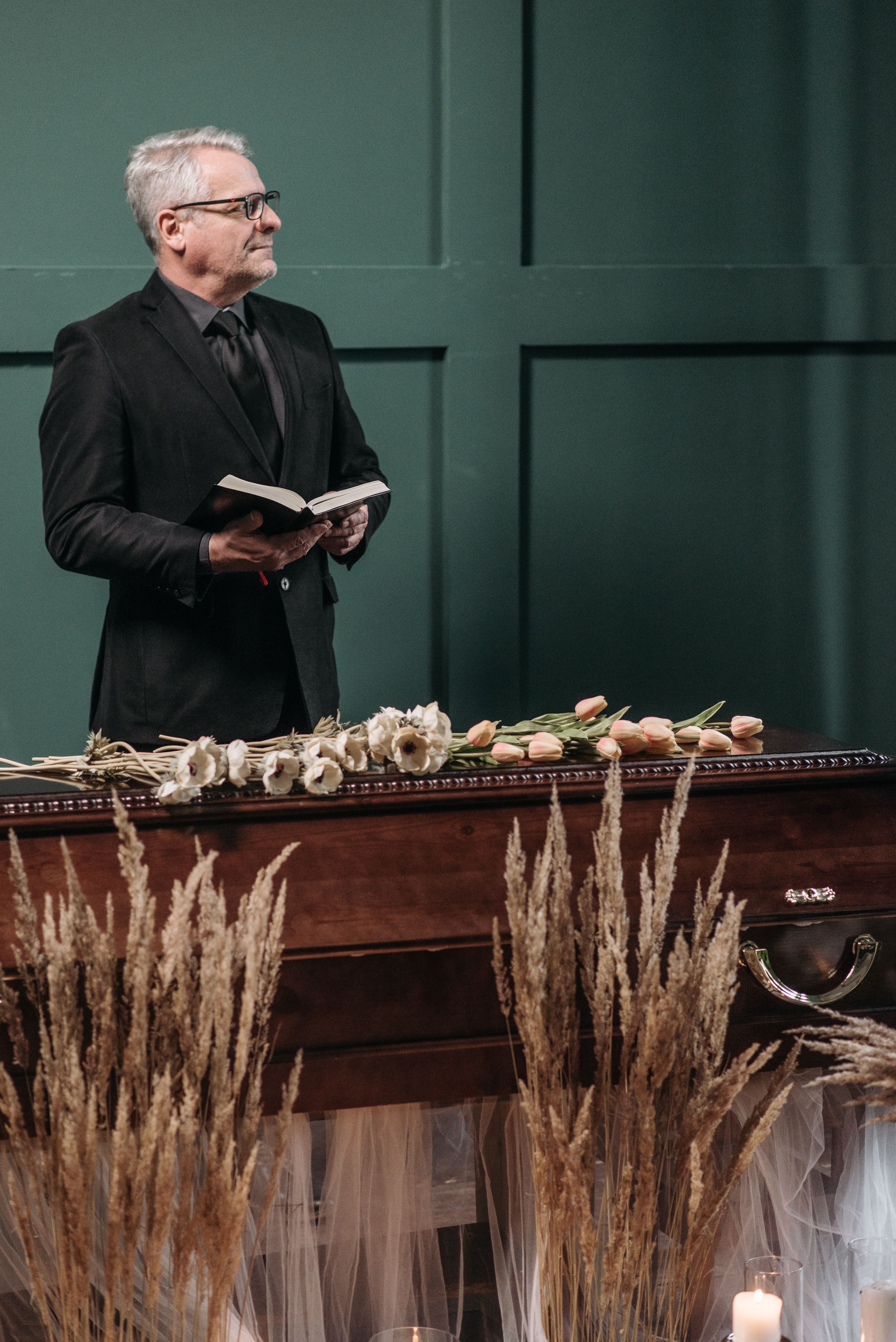 Mature man holding a book beside a coffin with flowers at a funeral service indoors.