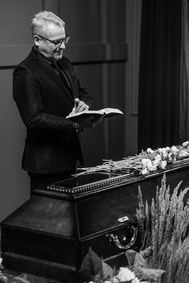 A Man Wearing Suit And Eyeglasses Reading A Book In Front Of A Wooden Coffin