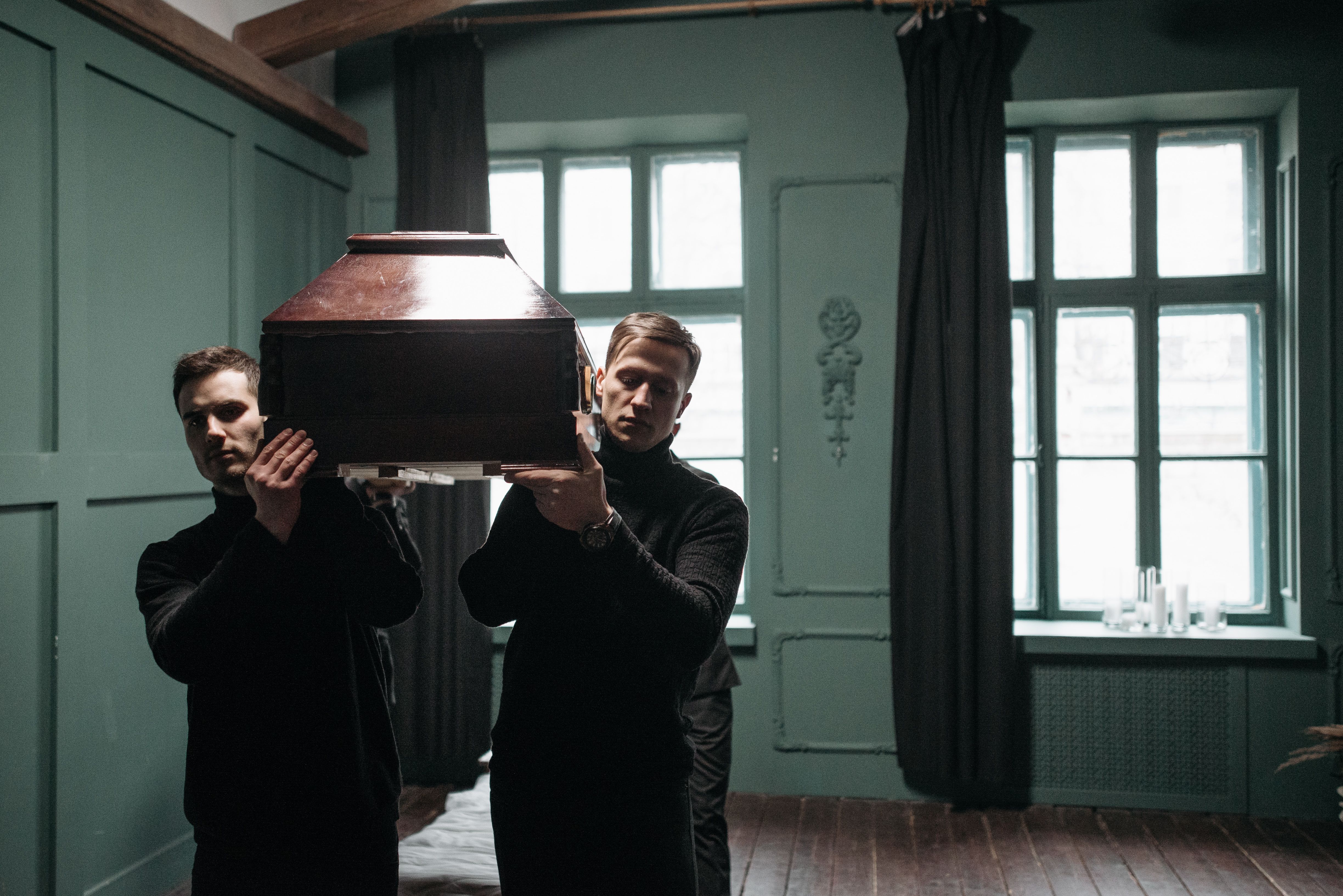 Two men in black attire carrying a wooden coffin through a dimly lit room.
