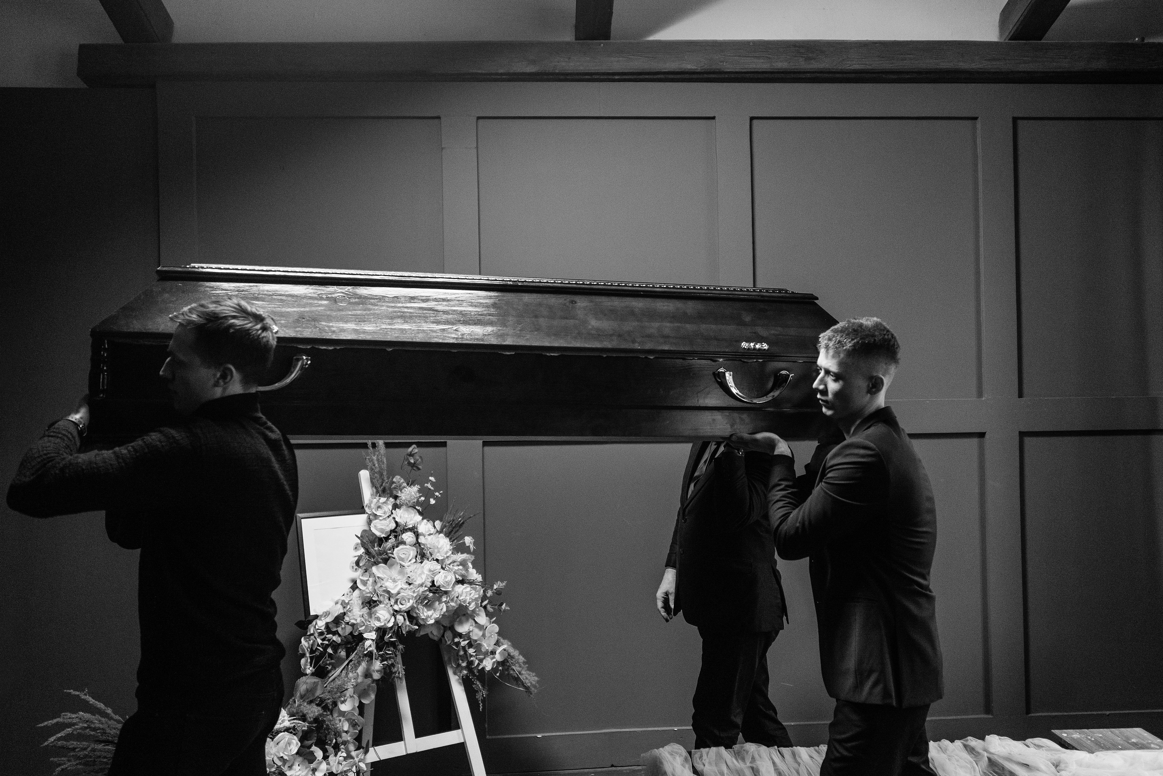 Men carrying a wooden coffin inside a funeral setting with flowers.