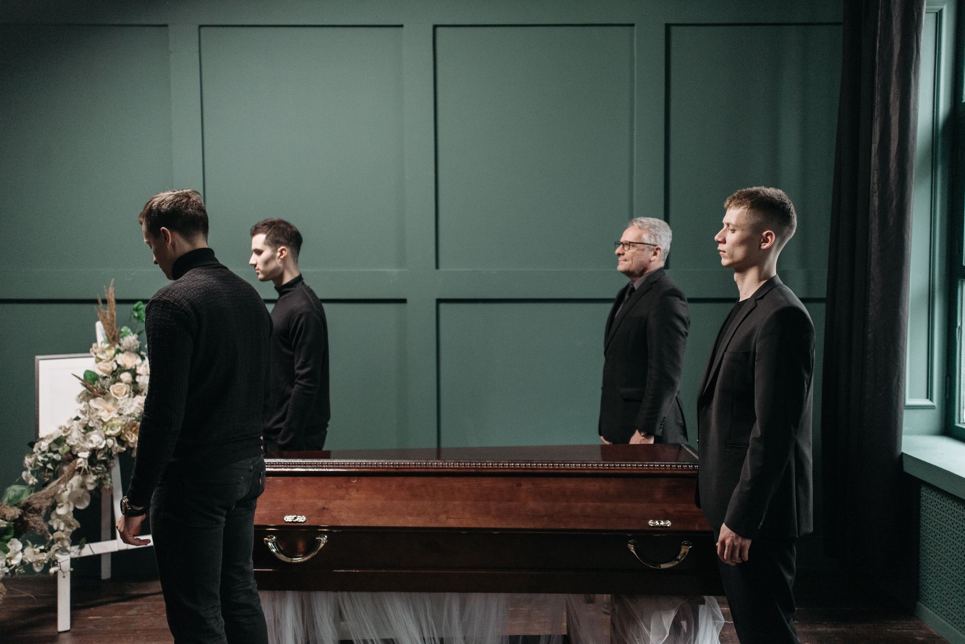 Four men in black suits standing at a funeral, symbolizing loss and mourning indoors.