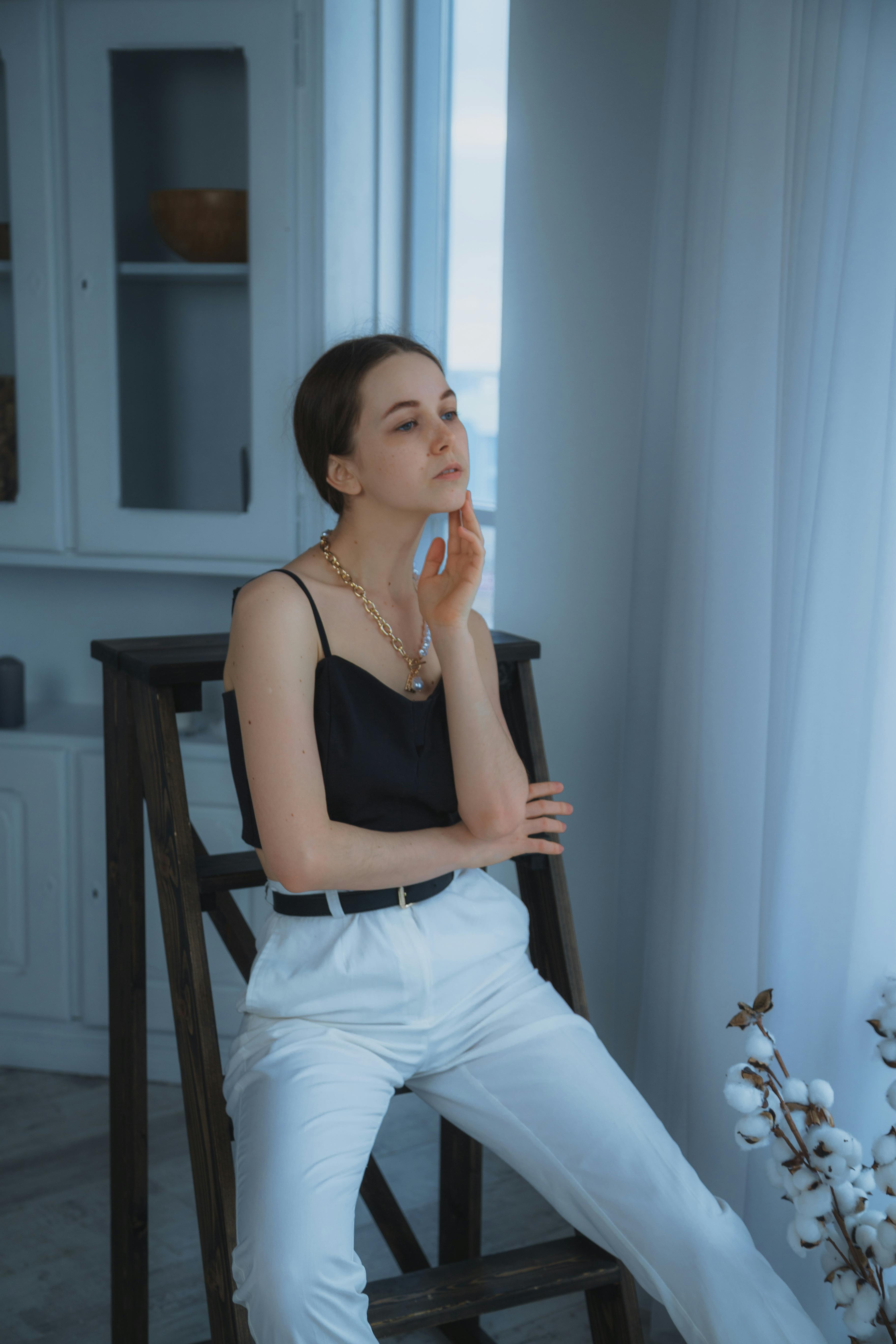 Pensive young female in stylish outfit looking away while sitting in light modern room near curtain and cupboards in apartment