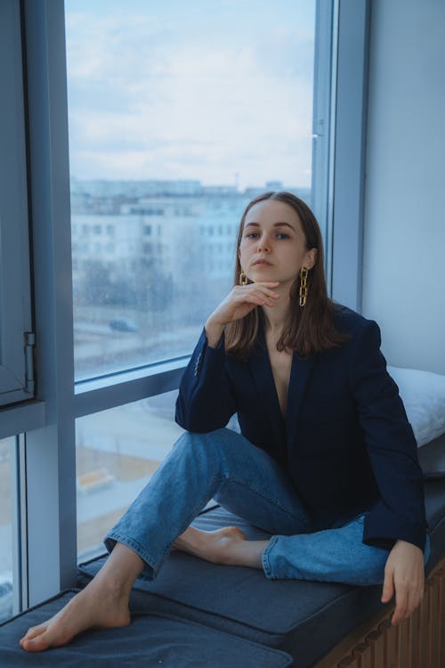 Attractive barefoot female wearing stylish outfit and earrings looking at camera with serious gaze while sitting near window in light room