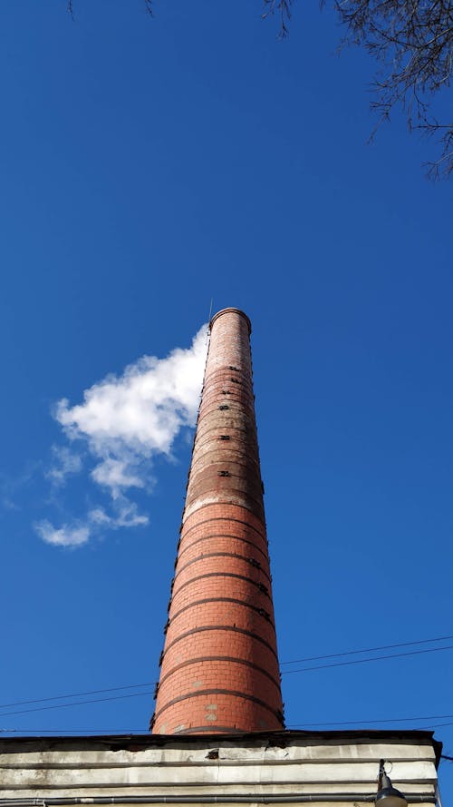 Low angle exterior of building with tall factory pipe with white smoke under blue cloudless sky in daytime