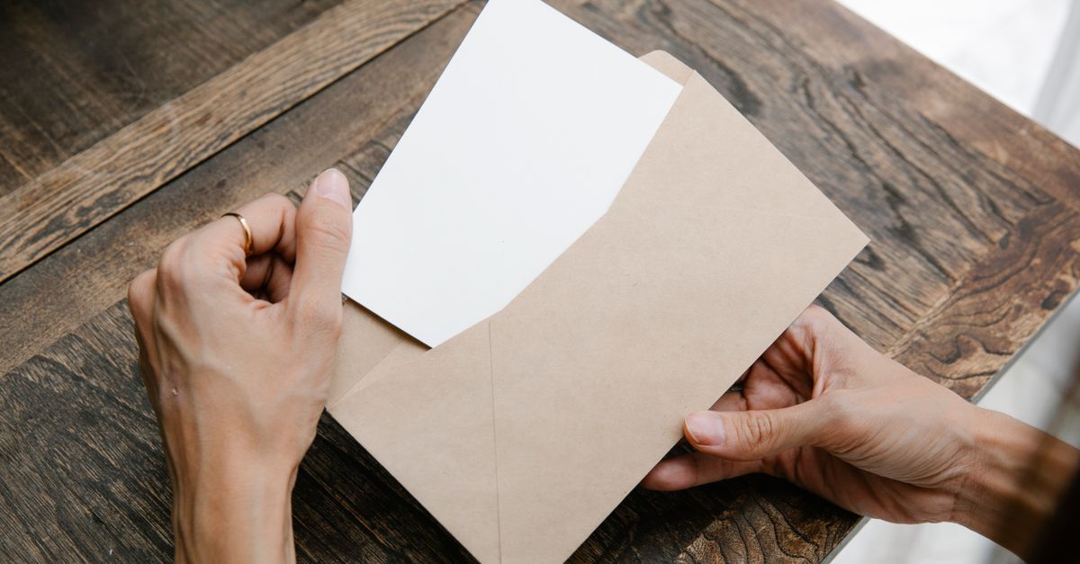Hands holding a letter inside a brown envelope on a wooden desk, viewed from above.