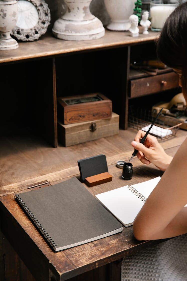 Woman Sitting At A Wooden Desk With A Blank Notebook Open In Front Of Her And Dipping A Pen In Ink 