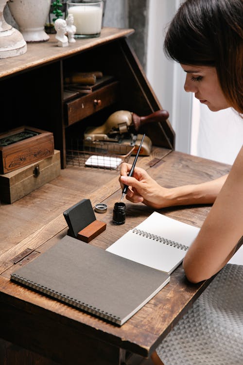 Woman Dipping Her Pen in Ink and Sitting at a Wooden Desk with Blank Notebook Pages in Front of Her 