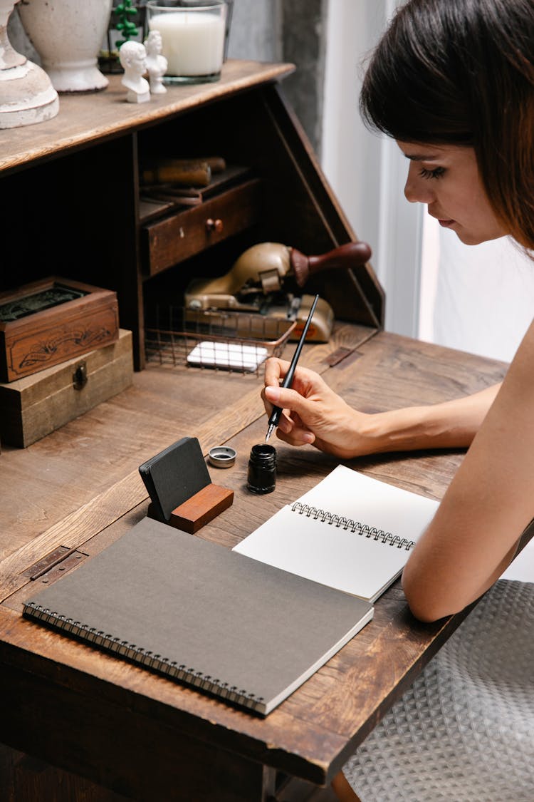 Woman Dipping Her Pen In Ink And Sitting At A Wooden Desk With Blank Notebook Pages In Front Of Her 