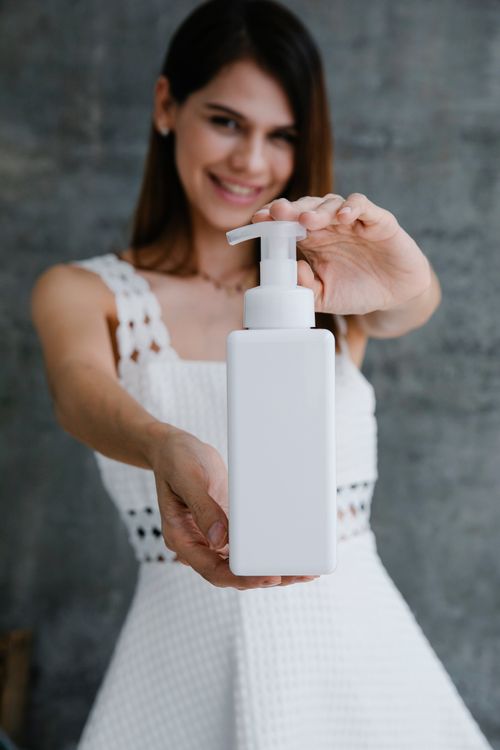 Free A cheerful woman in a white dress holding out a soap dispenser, focusing on hygiene. Stock Photo
