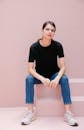 Young self assured woman in black t shirt and sneakers sitting on staircase while looking at camera on pastel background