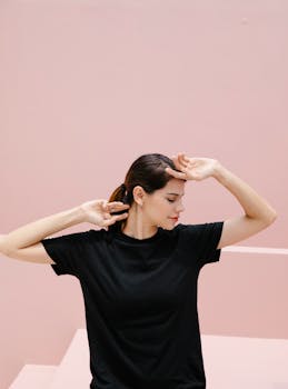 Young woman with brown hair in a black t-shirt strikes a reflective pose against a pastel pink backdrop.
