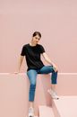 Millennial woman in sneakers and black t shirt looking at camera while sitting on fence in sunshine
