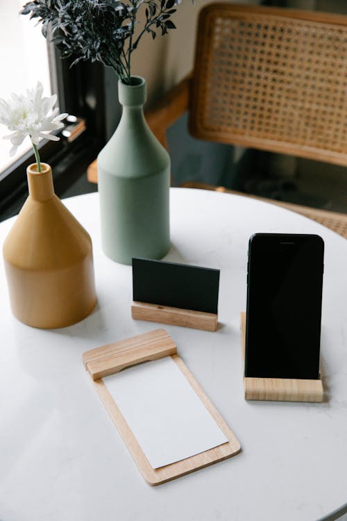 From above of wooden jotter with white sheets placed on round table near smartphone with black screen and visit card and composed with elegant vases with fresh flowers