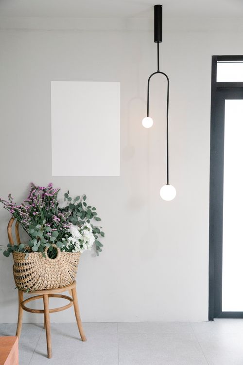 Free Stylish wicker bag with bunch of fresh aromatic flowers placed on wooden chair near wall with blank picture in light room with creative lamp Stock Photo