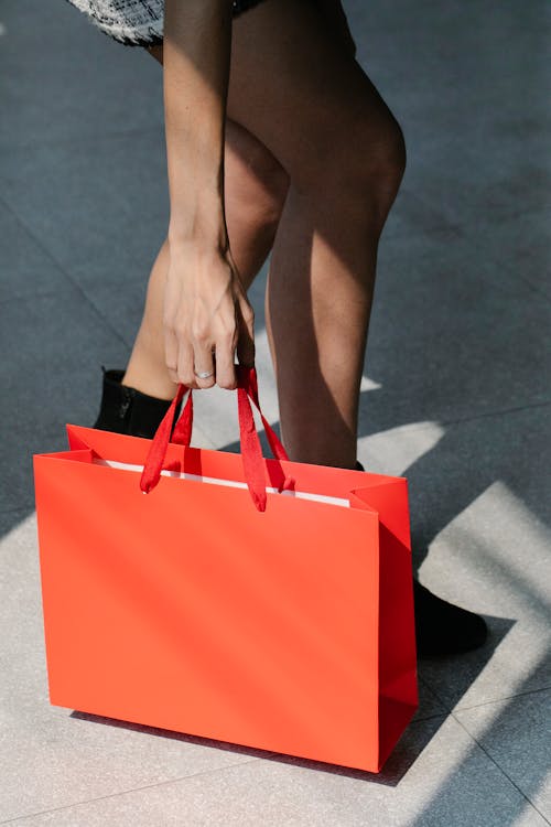 Free From above of crop faceless stylish female buyer in mini skirt and boots putting red paper bag on ground while standing on street after shopping Stock Photo