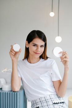 Self assured young female with long dark hair in white t shirt smiling while holding blank jars of cosmetic products in light room