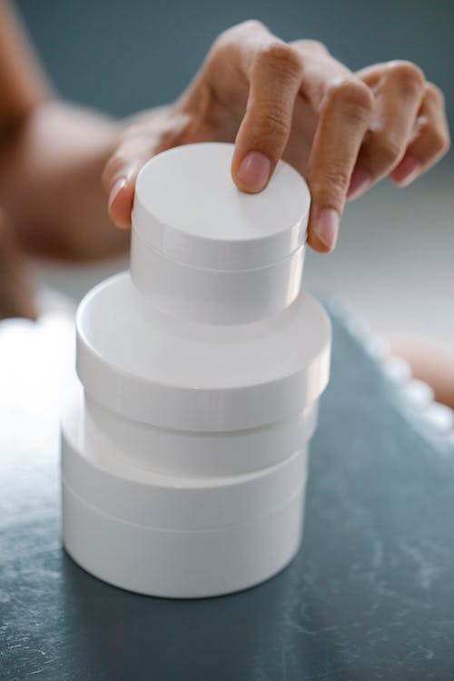 From above of crop anonymous lady stacking blank jars of different sizes of cosmetic products on table during skin care routine