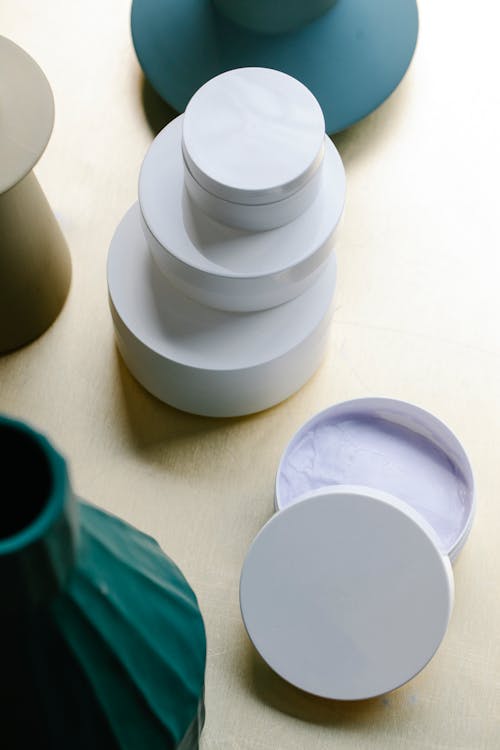 From above collection of various skin care products in minimalist white jars stacked on wooden table near vases in daylight