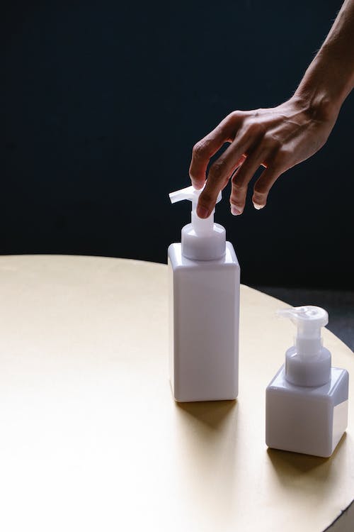 From above of crop anonymous female applying moisturizing cream from white bottle with dispenser placed on round table