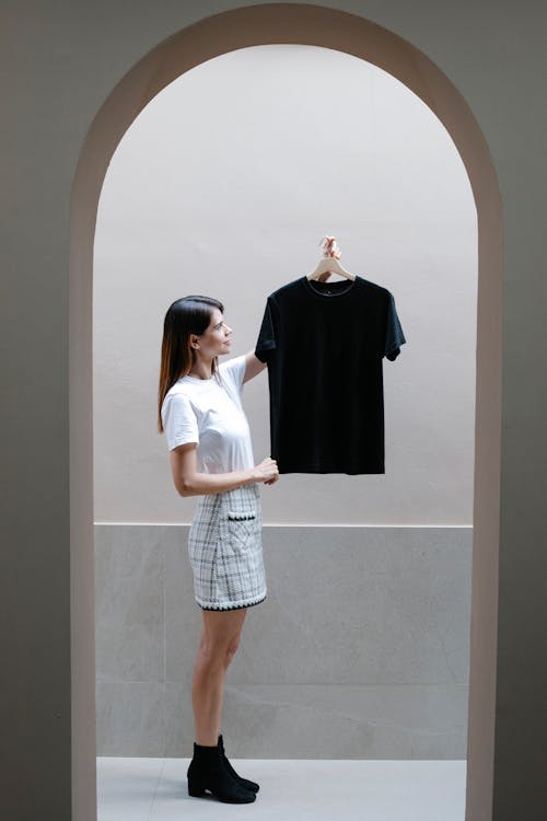 Side view full body of trendy young female designer in mini skirt and boots standing at arched doorway and demonstrating black t shirt