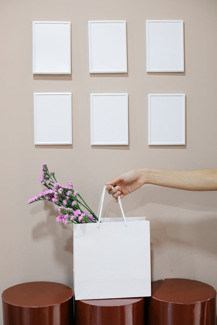 Crop Woman Holding Paper Bag With Flowers Near Wall With Blank Frames