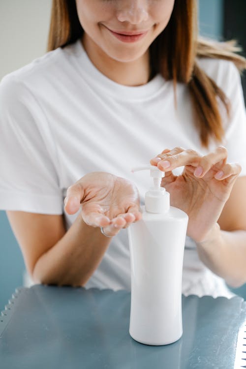 Crop unrecognizable happy female millennial in casual clothes smiling while applying moisturizing lotion on hand using dispenser of white bottle during skin care routine