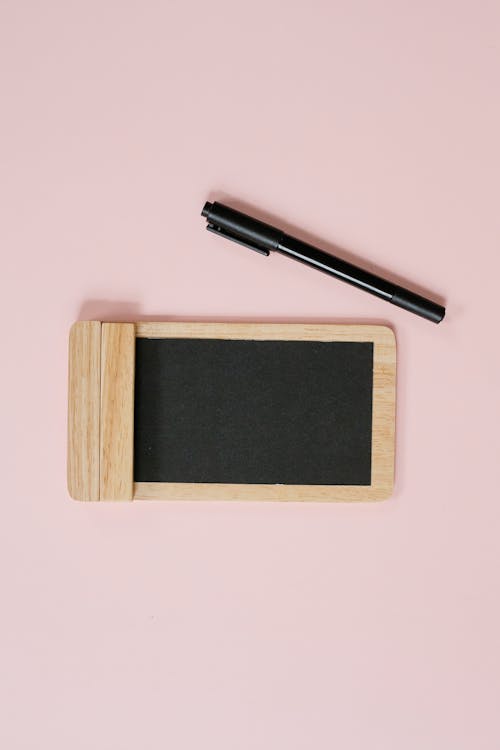 Overhead modern pen placed near timber notepad with empty black paper on pink background