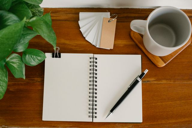 Open blank notebook, pen, mug and a trailing plant on a warm wooden table. Photo by Angela Roma on Pexels.