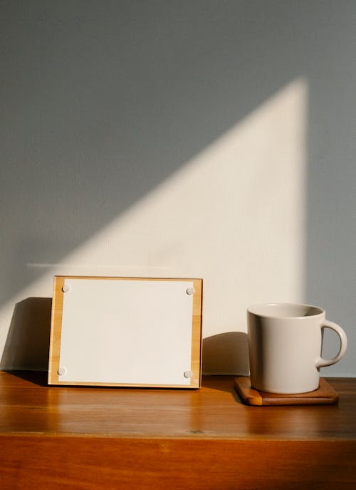 Cup of hot beverage and board with empty paper sheet placed on wooden table against gray wall in morning