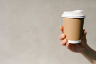 Anonymous woman demonstrating paper cup of takeaway coffee against gray background in sunny morning