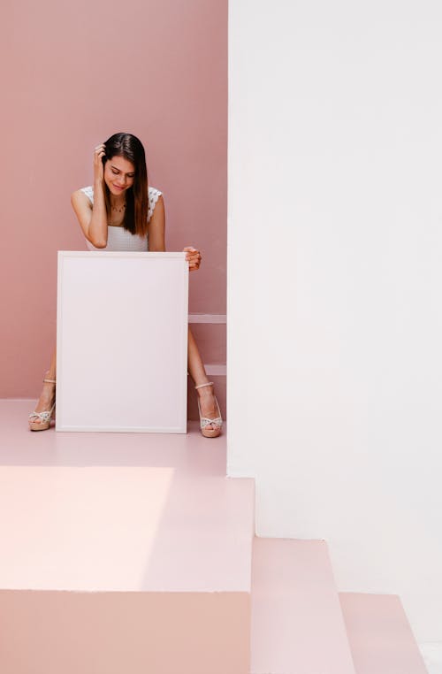 Free Stylish female touching hair and looking at empty poster while sitting near steps in pink room Stock Photo