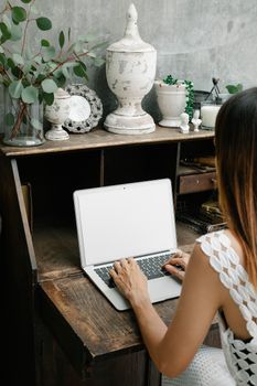 Back view unrecognizable female browsing modern netbook and sitting at old wooden desk decorated with vintage souvenirs