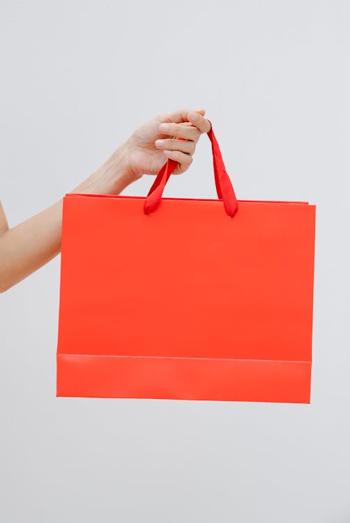 Free Crop unrecognizable female showing red paper shopping bag in hand against white wall Stock Photo