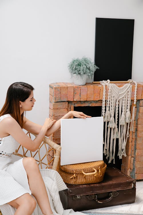 Free Trendy young female showing white blank canvas and sitting near brick fireplace in light room Stock Photo