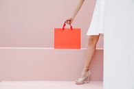 Crop anonymous stylish female in white dress and high heels carrying red shopping bag against pink wall