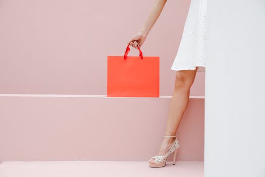 Crop anonymous stylish female in white dress and high heels carrying red shopping bag against pink wall