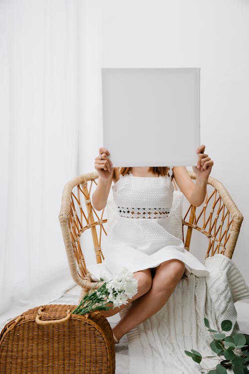 Unrecognizable female in stylish white dress showing square blank canvas and sitting on comfy wicker chair in light room