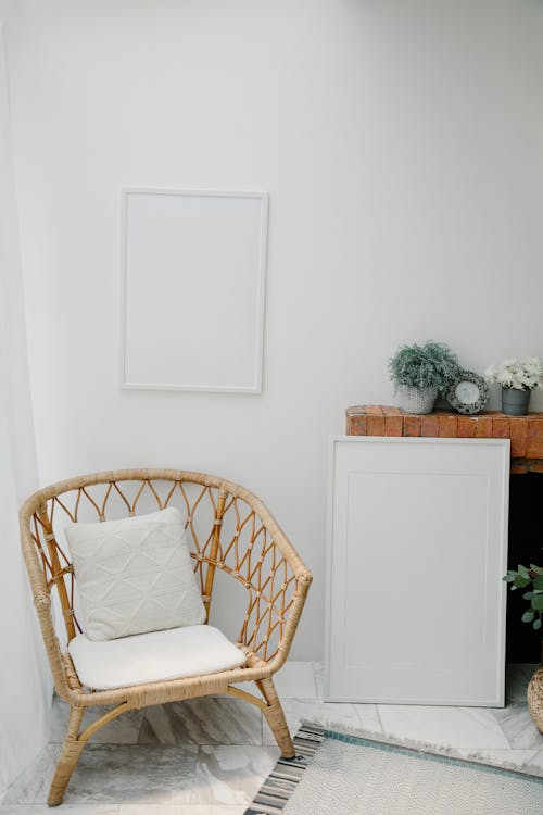 Free Comfortable armchair with cushions placed in corner under empty white frame hanging near shelf with potted plants Stock Photo