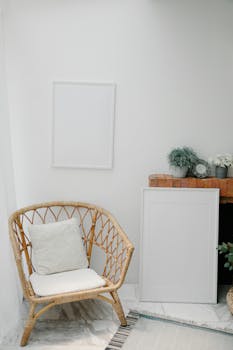 Comfortable armchair with cushions placed in corner under empty white frame hanging near shelf with potted plants