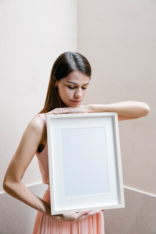 Young gentle female in pink dress showing white blank picture frame standing in corner against pink background