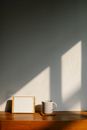 Frame with white empty picture and white mug of hot drink tea placed on wooden table at white wall in sunlight