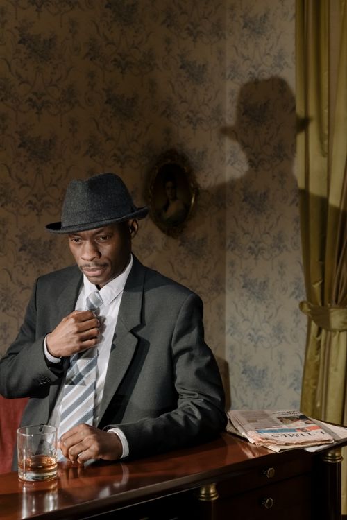 Free Black detective in a fedora hat with a drink in a vintage room setting, creating a noir mystery vibe. Stock Photo