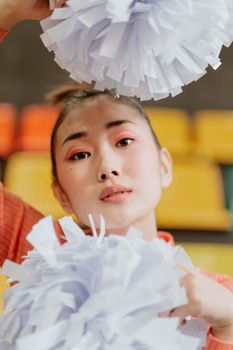 Close-up of an Asian cheerleader with colorful pom-poms indoors, showcasing a lively and energetic vibe.